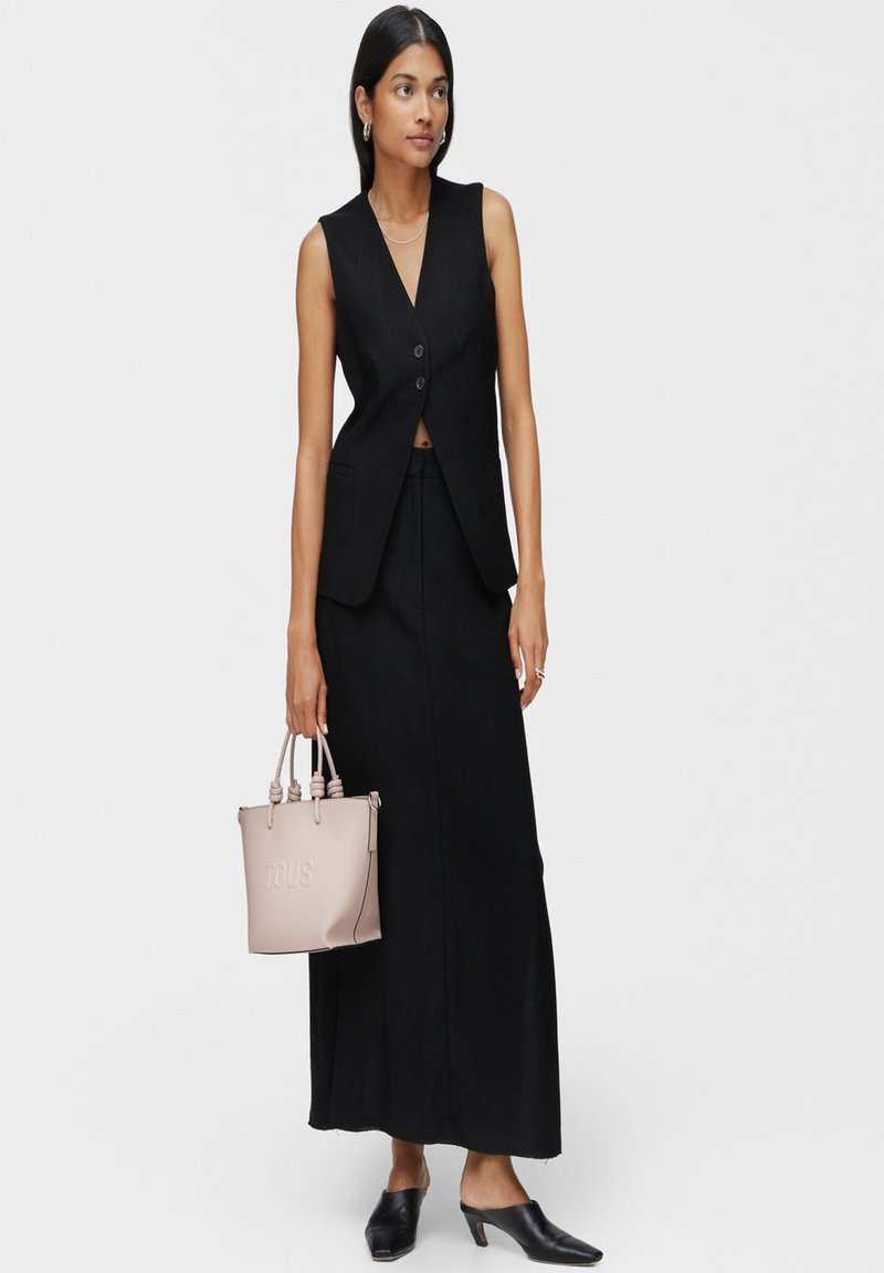 Chaleco negro sin mangas con escote en V profundo y botones delanteros, combinado con una falda larga negra. Los accesorios incluyen un bolso tote rosa claro.