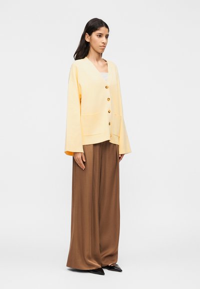 Γυναίκα που φοράει μια φαρδιά, ανοιχτό κίτρινη ζακέτα με κου μπιά, φαρδιά καφέ παντελόνια και μαύρα μυτερά παπούτσια, στέκεται μπροστά σε ένα απλό φόντο.