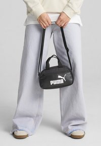 Borsa nera PUMA dalla forma arrotondata, con chiusura a zip e due manici. Presenta un logo bianco sul davanti, indossata in stile crossbody.