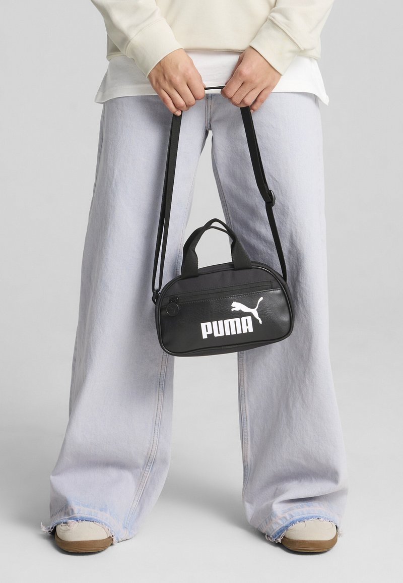 Borsa nera PUMA dalla forma arrotondata, con chiusura a zip e due manici. Presenta un logo bianco sul davanti, indossata in stile crossbody.