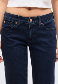 Άτομο που φοράει σκούρο μπλε τζιν με ψηλή μέση και χαλκομανιαστά κουμπιά και ένα κοντό λευκό τοπ που δείχνει γυμνή κοιλιά.