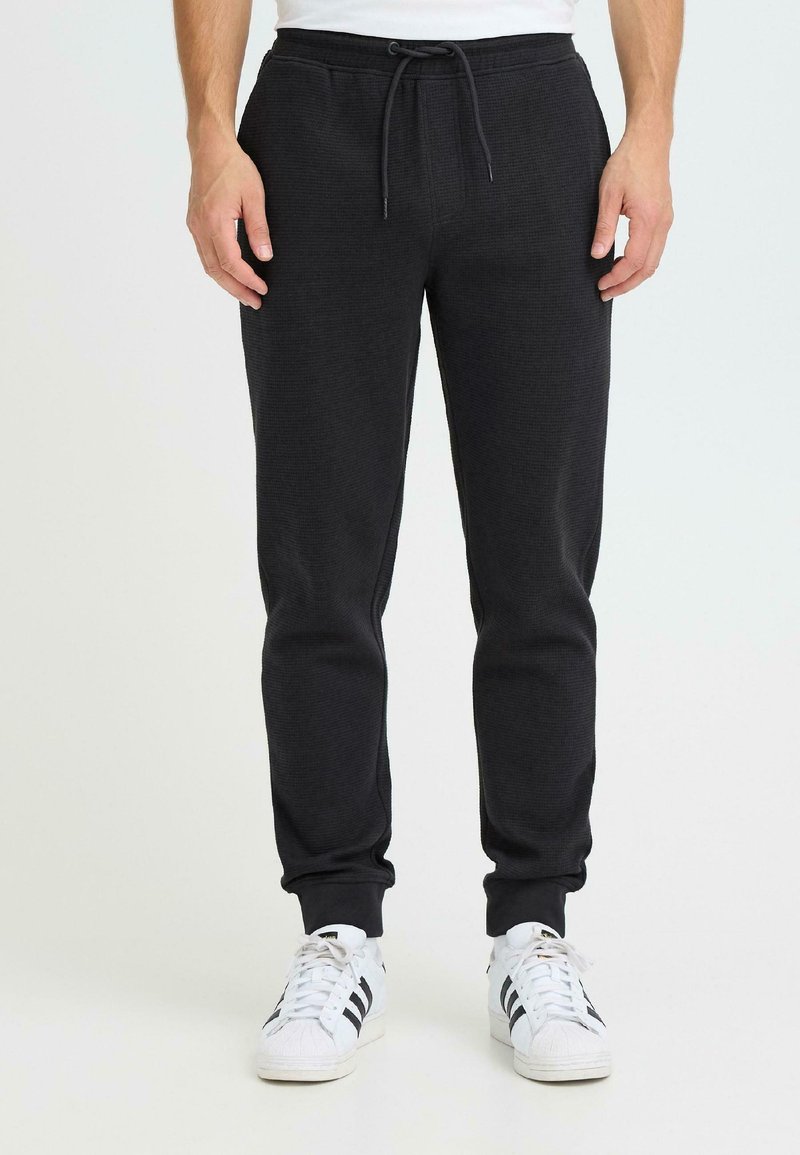 Czarne spodnie dresowe z teksturowanej tkaniny, z elastycznym pasem, regulowanym sznurkiem oraz ściągaczami przy kostkach, zestawione z białymi trampkami.