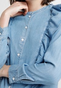 Camicia di jeans azzurro chiaro con scollo tondo, chiusura a bottoni e maniche arricciate, caratterizzata da una texture liscia e una vestibilità regolare.
