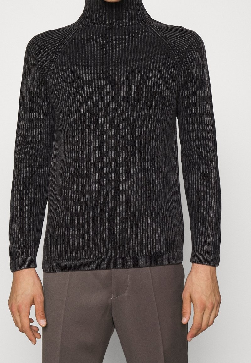 Pull à col roulé en tricot côtelé noir porté avec un pantalon marron ajusté, montrant le haut du corps et les bras sur un fond uni.