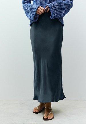 Personne portant une longue jupe en satin noire, un pull bleu en grosse maille, et des sandales plates marron à brides, debout contre un mur clair uni.