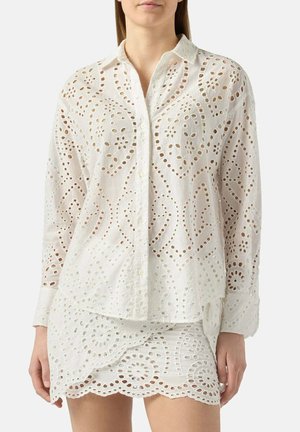 Vrouw draagt een wit blouse met lange mouwen van oogjeskant en een bijpassende minirok met bloemige en geometrische uitsnijdpatronen.