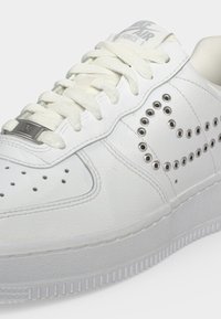 Άσπρο αθλητικό παπούτσι Nike Air Force 1 με διάτρητο σχέδιο swoosh από μεταλλικά οφθαλμιά και λευκά κορδόνια σε υφή σόλα.