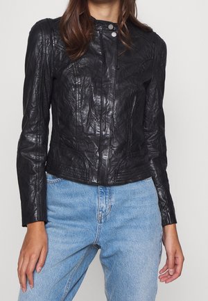 Woman wearing a fitted black leather jacket with snap buttons and light blue high-waisted jeans, standing against a plain background.