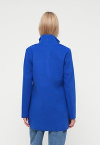Cappotto blu in misto lana con colletto alto, spalle strutturate e taglio dritto, visto di spalle su uno sfondo neutro.