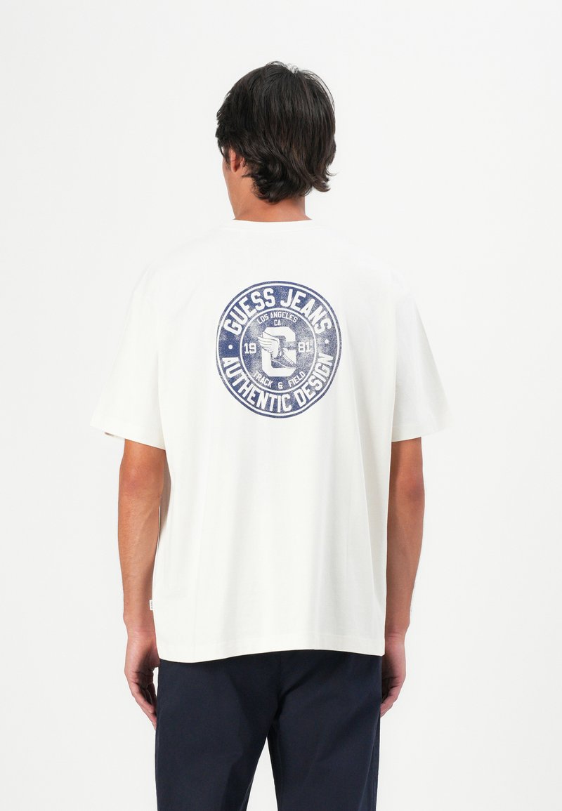 Mann mit dunklen Haaren, der ein weißes Guess Jeans T-Shirt mit einem blauen runden Logo auf dem Rücken und schwarze Hosen trägt, vor einem weißen Hintergrund stehend.