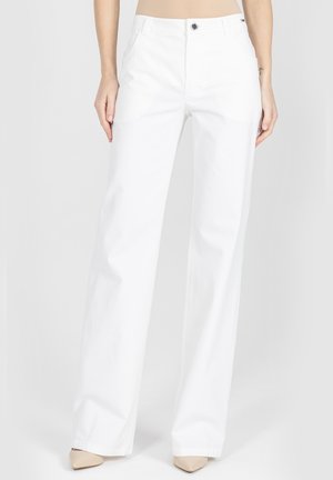 Kobieta ubrana w białe szerokie spodnie z wysokim stanem i beżowe szpilki z zaostrzonym noskiem, stojąca na jednolitym tle.