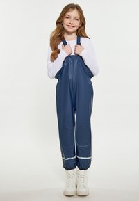 Námornícke modré nepremokavé overalové nohavice s nastaviteľnými popruhmi, textúrovaným povrchom, elastickými manžetami a kolennými záplatami. Nosené cez bielu košeľu s dlhým rukávom.