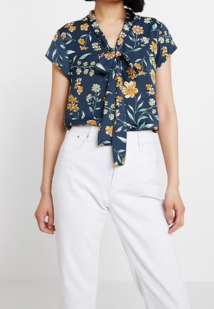 Blusa floral marina de manga corta, con detalle de lazo en la parte delantera, que presenta flores amarillas y verdes sobre una tela suave; combinada con pantalones blancos.
