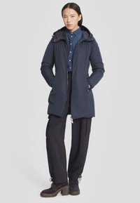 Marineblauwe gevoerde jas met een capuchon, ritssluiting aan de voorkant, zijzakken en elastische manchetten. Draag het over een blauwe overhemdd en zwarte broek met wijde pijpen.