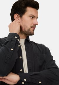 Chemise noire à boutons en tissu texturé, avec des boutons blancs, une poche poitrine et des manches longues. Portée par-dessus un t-shirt de couleur claire.