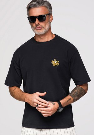 Homme aux cheveux gris et à la barbe portant des lunettes de soleil noires, un t-shirt noir avec un écusson abeille jaune, un pantalon blanc rayé et une montre-bracelet.