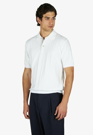 Uomo con polo bianca a maniche corte e pantaloni scuri, in piedi con le braccia rilassate, guarda verso sinistra su uno sfondo uniforme.