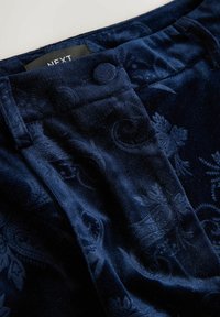 Dark blue velvet trousers with tone-on-tone paisley pattern, fabric-covered button, and visible brand label inside the waistband.