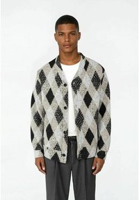 Man som bär en vit skjorta, grå byxor och en mönstrad cardigan med svarta och grå diamantformer, stående mot en helt vit bakgrund.