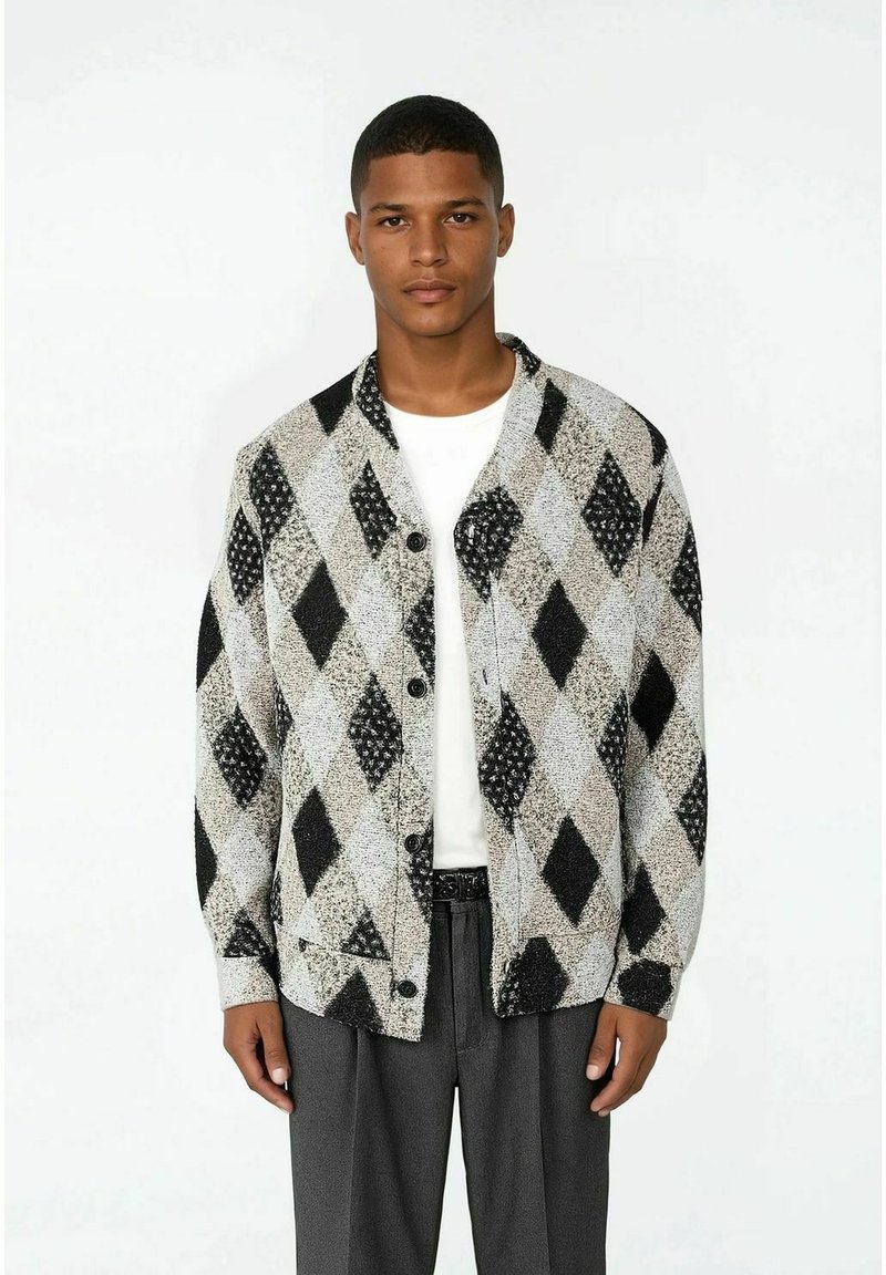 Man som bär en vit skjorta, grå byxor och en mönstrad cardigan med svarta och grå diamantformer, stående mot en helt vit bakgrund.