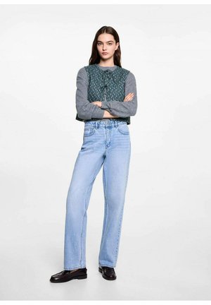 Groen geblokt cropped top met grijze mouwen, gedragen met lichtblauwe high-waisted jeans en zwarte schoenen. Model staat met de armen gekruist.