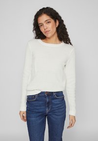 Pulover alb texturat din tricot, cu guler rotund și mâneci lungi, asortat cu blugi albaștri cu talie înaltă. Țesătura are un model subtil.