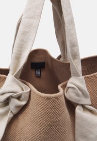 Sac fourre-tout en tissu brun texturé avec des poignées en coton beige, présentant des accents noués et une étiquette noire à l'intérieur.