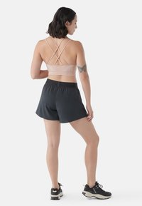 Sujetador deportivo de color rosa claro con tirantes entrecruzados, diseño texturizado; combinado con pantalones cortos deportivos negros y zapatillas de correr negras.
