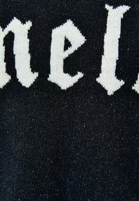 Acercamiento al tejido de punto negro con letras en estilo gótico blancas que muestran parcialmente la palabra "neil" tejida en el material.