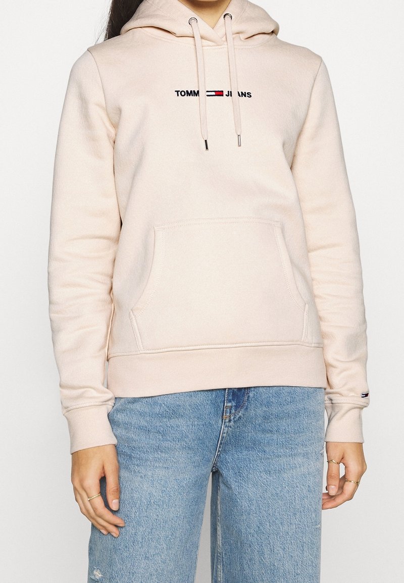 Personne portant un sweat à capuche beige clair avec le logo "Tommy Jeans" et un jean bleu, mains détendues le long du corps, cadré au niveau du cou et des cuisses.
