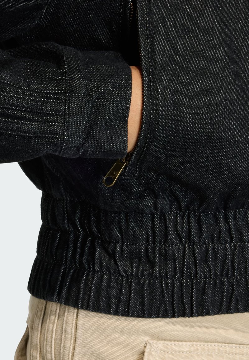 Veste en denim noir avec une taille froncée, dotée d'une poche zippée et de détails côtelés aux poignets, mettant en valeur une finition en tissu texturé.