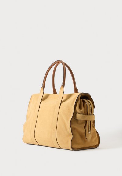 Sac à main en cuir souple couleur camel avec deux poignées arrondies marron et une poche zippée latérale, posé sur un fond clair uni.