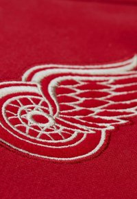 Roter Stoff mit einem weißen bestickten Design, das einem geflügelten Rad ähnelt, mit detaillierter Stickerei und einer glatten Textur.
