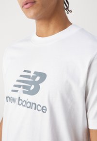 Bílá bavlněná tričko s velkým šedým vodorovným logem "NB" a textem "new balance" pod ním, s klasickým designem kulatého výstřihu.