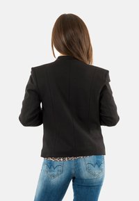 Femme aux longs cheveux bruns portant un blazer noir et un jean bleu, se tenant de dos contre un fond blanc uni.