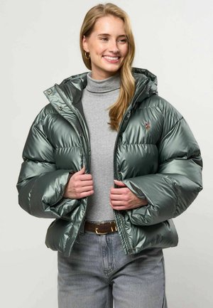 Femme souriante portant une doudoune verte brillante par-dessus un col roulé gris et un jean bleu clair avec une ceinture marron.