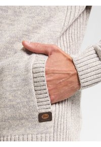 Vaaleanharmaa neulottu villapaita, jossa on ribatut hihansuut ja helma. Siinä on tasku, jossa on ruskea nahkainen logo-täg. Mukava rakenne ja yksityiskohtainen ommel.