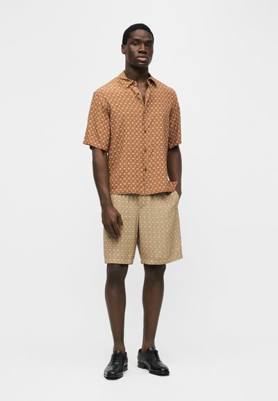 Homme debout portant une chemise à manches courtes marron à motifs, un short beige à motifs et des chaussures habillées noires, devant un fond clair uni.