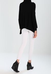 Schwarzer Pullover mit hohem Kragen und lockerer Silhouette, kombiniert mit schmalen weißen Hosen und schwarzen Ankle Boots mit Blockabsatz.