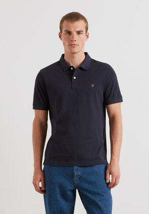 Giovane uomo in piedi che indossa una polo blu scuro tinta unita con un piccolo logo sul petto e jeans blu su sfondo bianco uniforme.