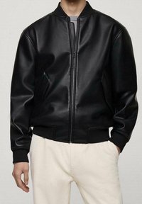 Homme portant une veste bomber en cuir noir avec fermeture éclair à l'avant et poignets côtelés, assortie à un pantalon de couleur claire, mains détendues le long du corps.