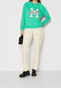 Mujer vistiendo un sudadera verde "Manhattan", pantalones de pierna ancha color crema, zapatos de plataforma negros y sosteniendo un pequeño bolso negro frente a un fondo blanco.