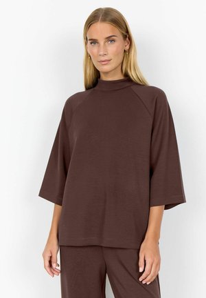 Donna con lunghi capelli biondi che indossa orecchini d'oro e un top marrone largo con maniche a tre quarti e pantaloni marroni abbinati.