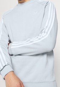 Světle modrý sweatshirt s kulatým výstřihem, který má bílé pruhy na rukávech a malé vyšívané logo na hrudi.