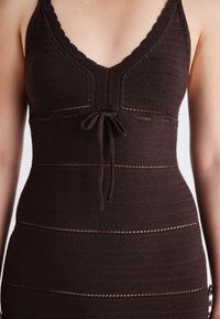 Ruda megzta suknelė su giliu V formos iškirpimu, banguotais dirželiais, rišimu priekyje ir horizontaliomis juostelėmis su permatomais akcentais.