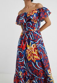 Maxi-jurk met off-shoulder flounces in blauwe, rode en gele gemusterde stof; heeft een ceintuur in de taille en rode franjerandaccenten.