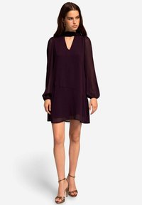 Paarse chiffon jurk met een diepe V-hals, lange opgezette mouwen en een gelaagde zoom. Gecombineerd met gouden, open schoenen met hoge hakken.