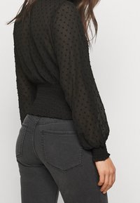 Top nero trasparente con punti testurizzati, maniche ampie e vita arricciata. Abbinato a jeans fitted grigi scuri con tasche posteriori.