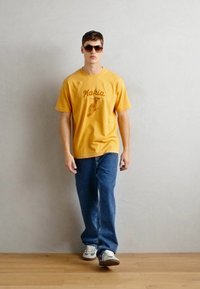 Gul bomull t-skjorte med brun grafisk trykk, blå denim jeans og grå joggesko. Modell iført mørke solbriller, står på tregulv.