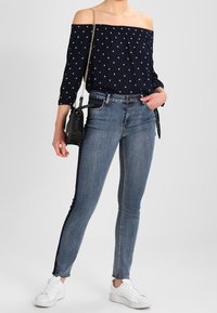 Blusa navy a spalle scoperte con pois bianchi, abbinata a jeans skinny in denim con strisce laterali e sneakers bianche. Inclusa una borsa nera.